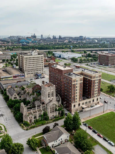 Aerial view of an abandoned, ivy-covered stone church ruin next to a tall red brick hotel and other downtown Gary buildings, with a steel mill in the distance.
